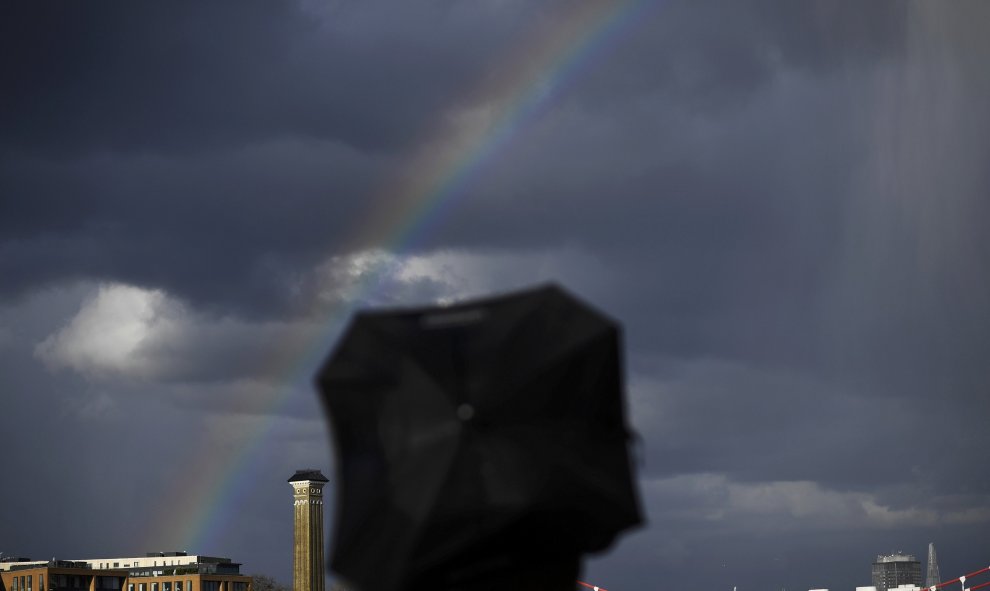 Un hombre se refugia de la lluvia mientras camina con un arco iris al fondo en Londres. REUTERS/Dylan Martinez