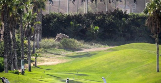 Una docena de subsaharianos permanece encaramada a la valla de Melilla, mientras dos personas juegan al golg. José Palazón tituló esta imagen como ‘Paisajes de desolación’.