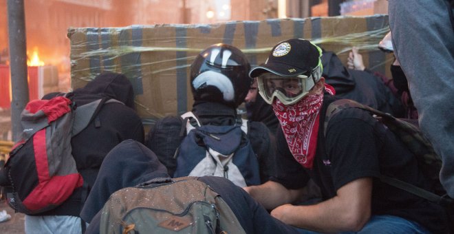 Varios jóvenes encapuchados se protegen de las balas de goma con un parapeto de cartón y cinta de embalaje, durante los disturbios de Barcelona. GUILLEM SANS