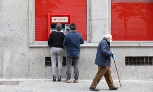 Un hombre con bastón camina por la capital al lado de dos personas que sacan dinero de un cajero, en Madrid, a 28 de abril de 2020.